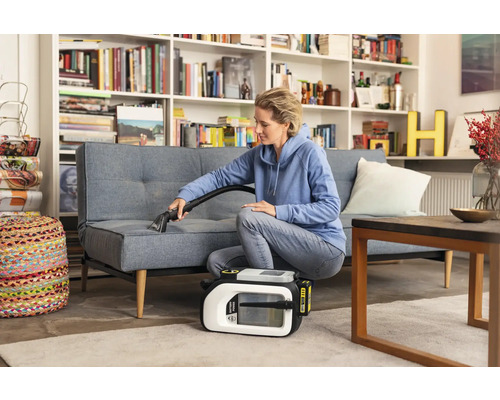 Frau reinigt Sofa mit einem Waschsauger in einem Wohnzimmer mit Bücherregal