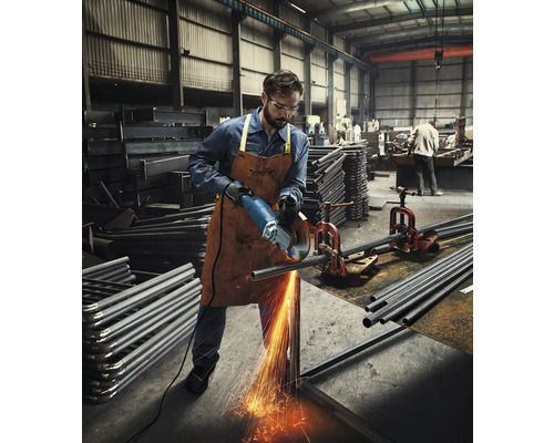 Un homme coupe un tube métallique avec une meuleuse dans un atelier, produisant des étincelles.
