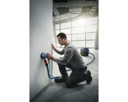 Un homme ponce un mur avec une ponceuse murale raccordée à un aspirateur.