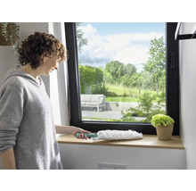 Frau wischt Fensterbrett mit Staubwedel. Fenster mit Blick ins Grüne.