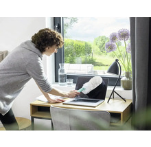 Frau reinigt Laptop mit Staubwedel auf Schreibtisch.