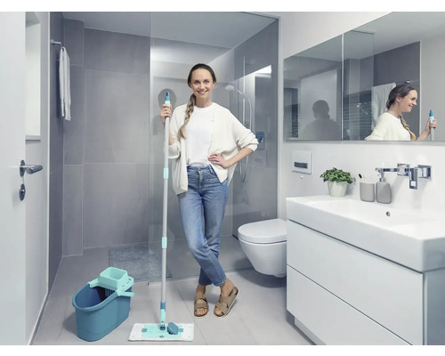 Helles Badezimmer mit Frau, die einen Mopp und einen Eimer hält, neben einem Waschbeckenunterschrank, einer Toilette und einer Dusche.