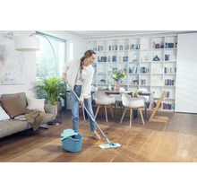 Frau wischt Holzboden mit Mopp und Eimer in einem hellen Wohnzimmer.