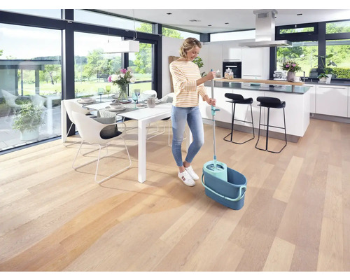 Frau wischt Holzboden mit Wischmop und Eimer in einem hellen Wohnbereich.