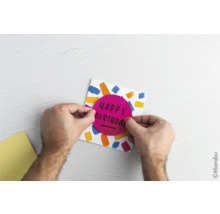 "Eine Person gestaltet eine Geburtstagskarte mit bunten Farben und einem runden Aufkleber mit der Aufschrift ""Happy Birthday""."