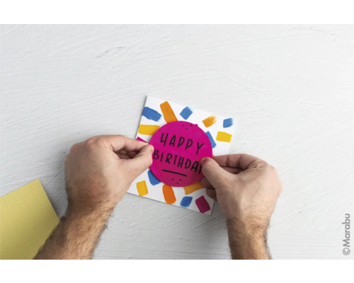 "Eine Person gestaltet eine Geburtstagskarte mit bunten Farben und einem runden Aufkleber mit der Aufschrift ""Happy Birthday""."