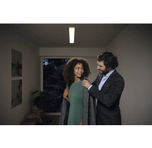 Szene mit einer Frau, die einen Mantel anprobiert, und einem Mann in einem formellen Outfit, beleuchtet durch eine Deckenleuchte.