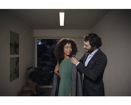 Szene mit einer Frau, die einen Mantel anprobiert, und einem Mann in einem formellen Outfit, beleuchtet durch eine Deckenleuchte.