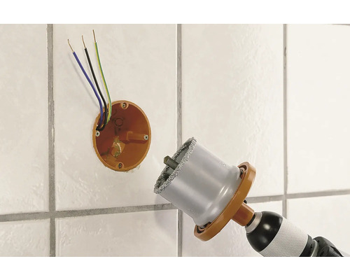 Scie cloche diamantée perçant un trou dans un mur carrelé à côté d'une boîte de dérivation avec des câbles électriques.