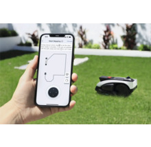 Une personne tient un smartphone avec une application pour contrôler un robot tondeuse. À l'arrière-plan, le robot tondeuse se trouve sur une pelouse.