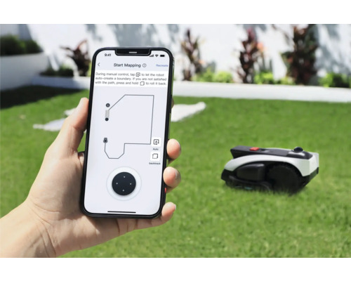 Une personne tient un smartphone avec une application pour contrôler un robot tondeuse. À l'arrière-plan, le robot tondeuse se trouve sur une pelouse.