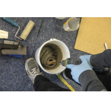 Mélange de colle à carrelage dans un seau avec un agitateur électrique. Des outils et une personne portant des gants de protection sont également visibles.