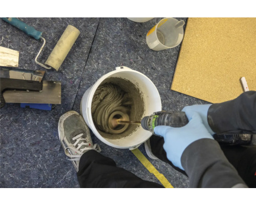 Mélange de colle à carrelage dans un seau avec un agitateur électrique. Des outils et une personne portant des gants de protection sont également visibles.