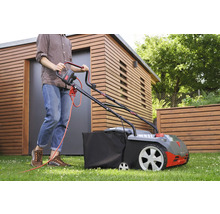 Eine Person benutzt einen Rasenmäher auf einer Rasenfläche vor einem Holzhaus.