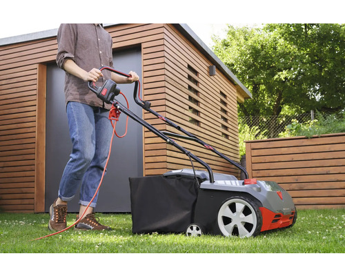 Eine Person benutzt einen Rasenmäher auf einer Rasenfläche vor einem Holzhaus.