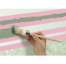 Une personne peint une ligne verte avec un pinceau rond sur un mur avec des rayures roses et blanches