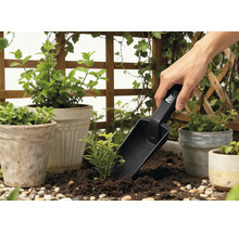 Scène de jardin avec pots de plantes, verdure environnante et une personne utilisant une transplanteuse.