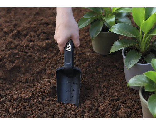 Une main tient une pelle à planter dans la terre, à côté de plantes en pot.