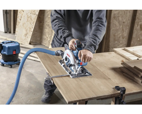Une personne scie une planche de bois avec une scie circulaire portative, un aspirateur est branché.