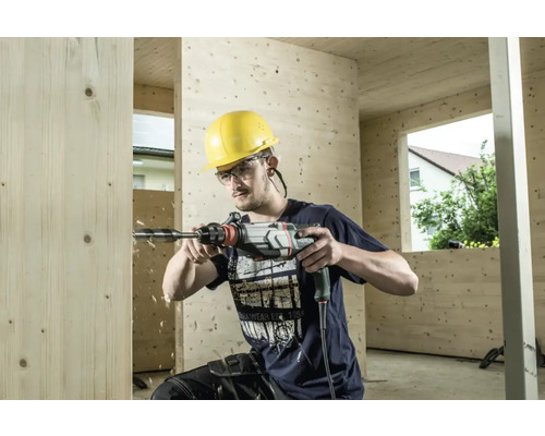 Un homme perce un trou dans une paroi en bois avec une perceuse. Il porte un casque de protection jaune et des lunettes de sécurité.