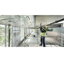 Un ouvrier du bâtiment, portant un casque de protection et un gilet de sécurité, utilise une perceuse sur un chantier pour fixer un chemin de câbles au plafond.