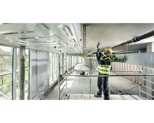 Un ouvrier du bâtiment, portant un casque de protection et un gilet de sécurité, utilise une perceuse sur un chantier pour fixer un chemin de câbles au plafond.