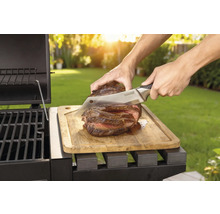 Un steak cuit est coupé au couteau sur une planche à découper en bois à côté d'un barbecue.