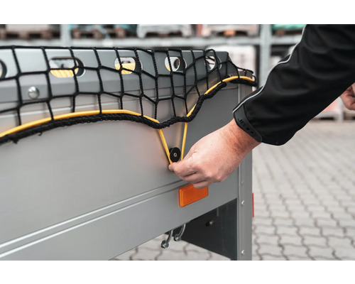 Hand befestigt ein Anhängernetz mit einer elastischen Schnur an einem Befestigungsknopf an der Bordwand eines Pkw-Anhängers.