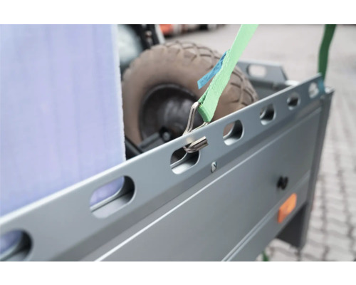 Nahaufnahme eines grünen Spanngurts mit Metallhaken, der an der Seitenreling eines Metallanhängers befestigt ist.