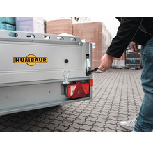 Eine Person öffnet den Bordwandverschluss eines silbernen Humbaur Anhängers mit Humbaur Logo.