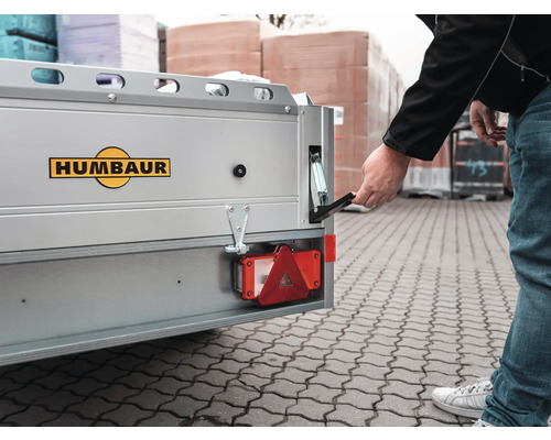 Eine Person öffnet den Bordwandverschluss eines silbernen Humbaur Anhängers mit Humbaur Logo.