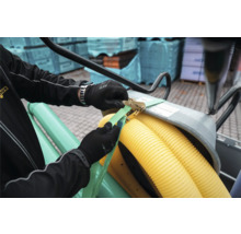 Eine Person mit Arbeitshandschuhen sichert Wellrohre aus Kunststoff mit einem Spanngurt und einer Metallratsche.