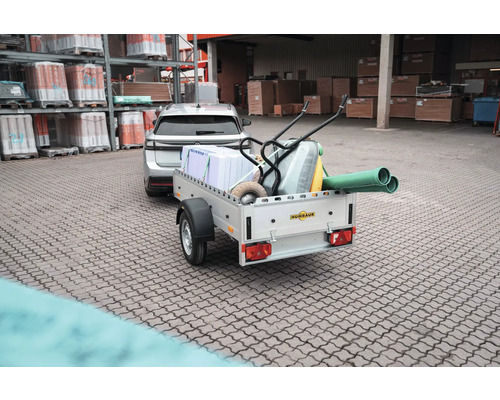 Beladener Humbaur Anhänger mit Dämmplatten, Schubkarre und Rohren, angekuppelt an ein Auto vor einem Baustofflager.