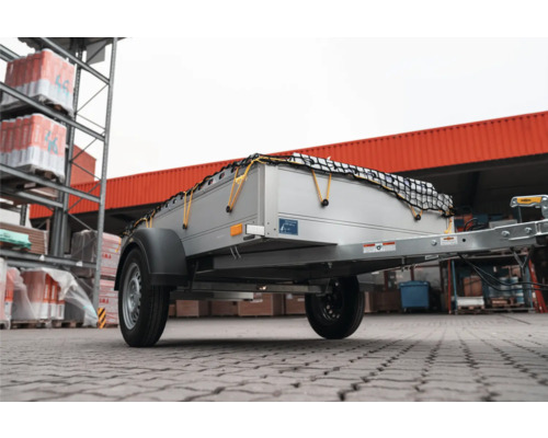 Einachsiger Anhänger für Autos mit silberner Ladefläche und schwarzem Ladungssicherungsnetz im Außenbereich eines Baumarktes.