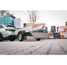 Anhänger an einem Auto befestigt, beladen mit einer verpackten Rolle auf einem Lagergelände mit Baumaterialien.