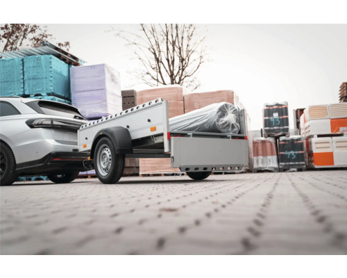 Anhänger an einem Auto befestigt, beladen mit einer verpackten Rolle auf einem Lagergelände mit Baumaterialien.