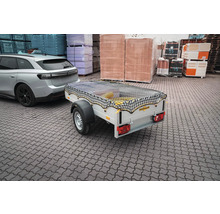 Humbaur PKW-Anhänger mit schwarzem Ladungssicherungsnetz, angekoppelt an einen silbernen PKW auf einem Lagerplatz mit Baumaterialien.