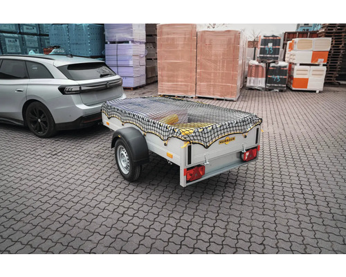 Humbaur PKW-Anhänger mit schwarzem Ladungssicherungsnetz, angekoppelt an einen silbernen PKW auf einem Lagerplatz mit Baumaterialien.