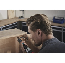 Un homme fixe une charnière à un meuble en bois avec une visseuse sans fil.