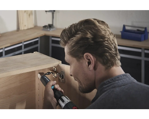 Un homme fixe une charnière à un meuble en bois avec une visseuse sans fil.