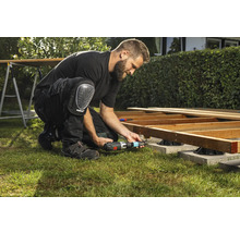 Un homme construit une terrasse en bois avec une visseuse sans fil et des genouillères dans le jardin.