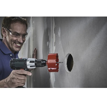 Un homme portant des lunettes de sécurité perce un trou dans un mur à l'aide d'une scie cloche.