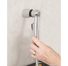 Une main tient une douchette de bidet avec un flexible de douche sur un support mural dans la salle de bains.