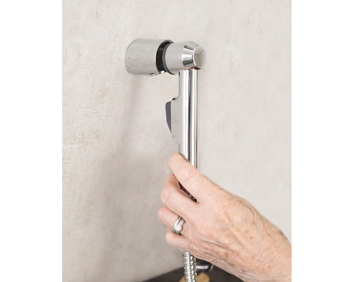 Une main tient une douchette de bidet avec un flexible de douche sur un support mural dans la salle de bains.