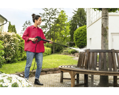 Frau reinigt eine runde Holzbank mit einem Hochdruckreiniger im Garten.