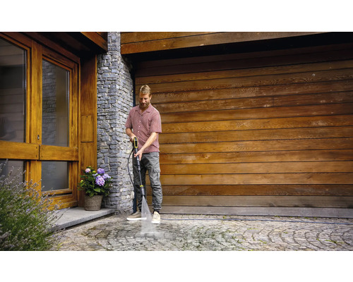 Ein Mann reinigt einen gepflasterten Weg mit einem Hochdruckreiniger vor einer Garage.