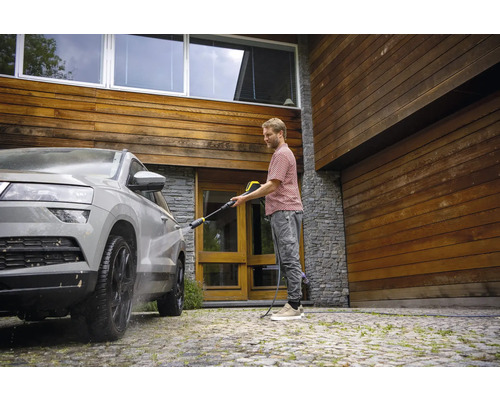 Mann reinigt Auto mit Hochdruckreiniger vor einem Haus mit Holzfassade