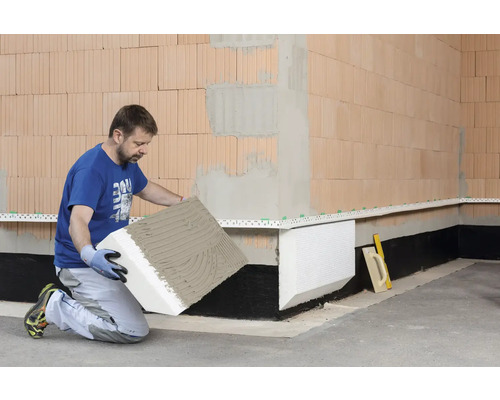 Mann verlegt Perimeterdämmplatten an einer Hauswand aus Ziegelsteinen.