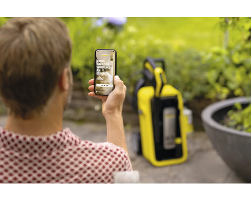 Eine Person hält ein Smartphone mit der Kärcher-App in der Hand, im Hintergrund ein Kärcher Hochdruckreiniger.