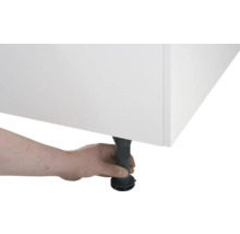 Une main ajuste un pied de meuble noir réglable en hauteur sous un meuble blanc.